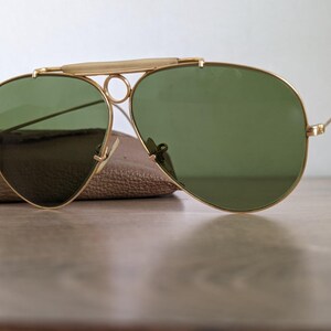 K&ouml;nnte beinhalten: Goldfarbene Pilotenbrille mit gr&uuml;nen Gl&auml;sern. Die Sonnenbrille hat einen Doppelsteg und einen Brauenb&uuml;gel. Die Gl&auml;ser sind tropfenf&ouml;rmig. Die Sonnenbrille liegt auf einem braunen Lederetui.