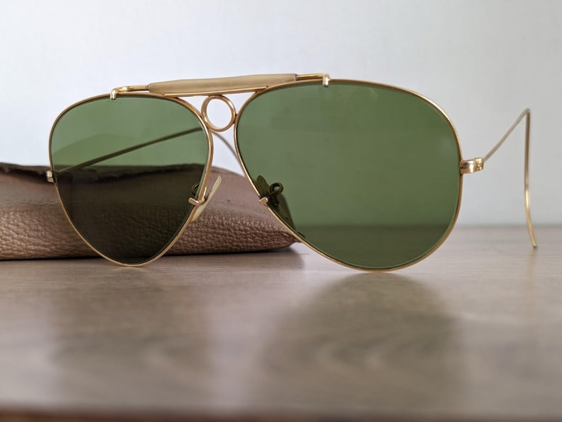 K&ouml;nnte beinhalten: Goldfarbene Pilotenbrille mit gr&uuml;nen Gl&auml;sern. Die Sonnenbrille hat einen Doppelsteg und einen Brauenb&uuml;gel. Die Gl&auml;ser sind tropfenf&ouml;rmig. Die Sonnenbrille liegt auf einem braunen Lederetui.