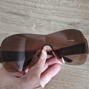 May include: Brown oversized sunglasses with a curved design. The sunglasses have a dark brown tint and a thin, curved frame. The arms of the sunglasses are a darker brown color. The sunglasses are held by a hand.