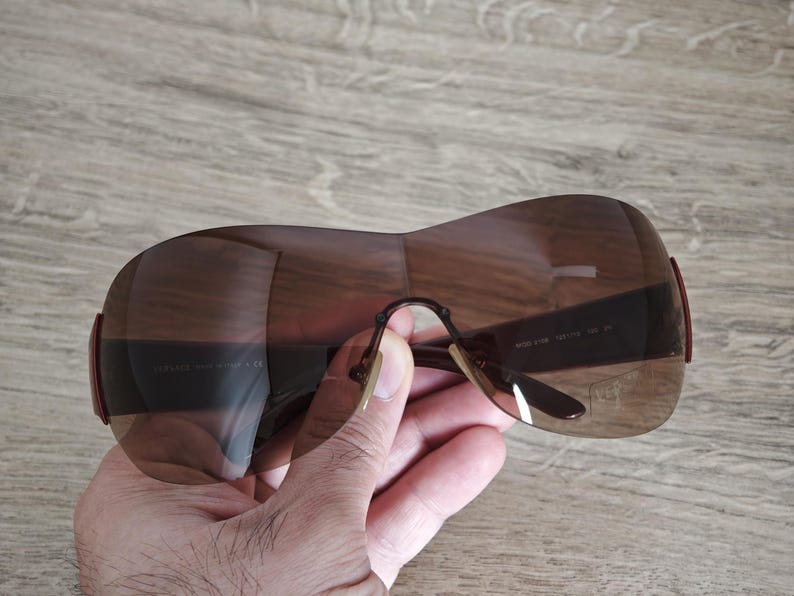 May include: Brown oversized sunglasses with a curved design. The sunglasses have a dark brown tint and a thin, curved frame. The arms of the sunglasses are a darker brown color. The sunglasses are held by a hand.