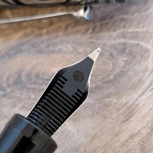 以下が含まれることがあります： 数字の「4」が見える黒い万年筆のペン先のクローズアップ。ペン先は、銀色のペンバレルと木製の表面のぼやけた背景に対して設定されています。ペンは書くように設計されています。