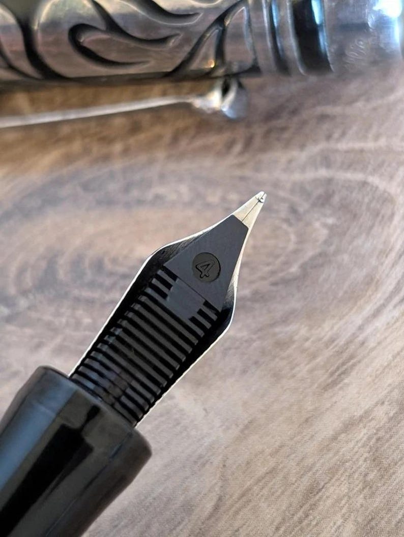 以下が含まれることがあります： 数字の「4」が見える黒い万年筆のペン先のクローズアップ。ペン先は、銀色のペンバレルと木製の表面のぼやけた背景に対して設定されています。ペンは書くように設計されています。