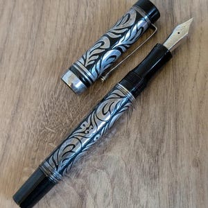 以下が含まれることがあります： 黒とシルバーの万年筆。装飾的なフローラルデザインが施されています。 ペンのキャップは外されており、ペン先が見えています。 ペンにはシルバーのクリップが付いており、木製の背景に置かれています。 装飾的なデザインはキャップにも繰り返されています。