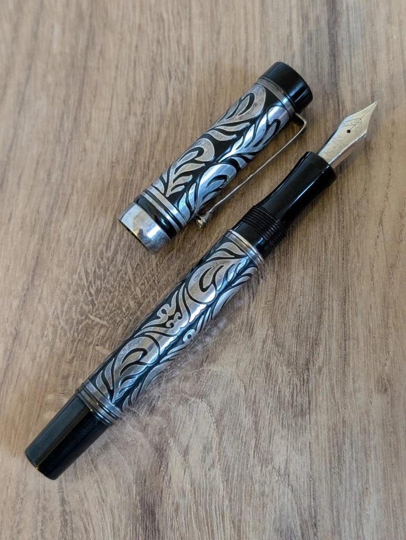 以下が含まれることがあります： 黒とシルバーの万年筆。装飾的なフローラルデザインが施されています。 ペンのキャップは外されており、ペン先が見えています。 ペンにはシルバーのクリップが付いており、木製の背景に置かれています。 装飾的なデザインはキャップにも繰り返されています。