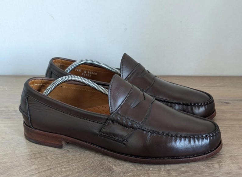 May include: A pair of brown leather penny loafers. The shoes have a classic design with a rounded toe, a strap across the vamp, and a low heel. The leather has a polished finish.
