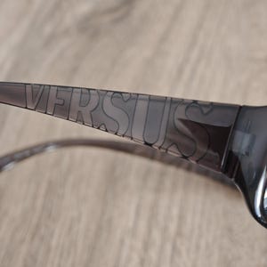 Puede incluir: Un par de gafas de sol de color gris oscuro transl&uacute;cido con la palabra VERSUS grabada en letras may&uacute;sculas grandes a lo largo de la patilla. La montura est&aacute; fabricada con un material pl&aacute;stico liso y semitransparente con acabado brillante.