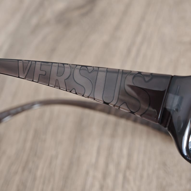 Puede incluir: Un par de gafas de sol de color gris oscuro transl&uacute;cido con la palabra VERSUS grabada en letras may&uacute;sculas grandes a lo largo de la patilla. La montura est&aacute; fabricada con un material pl&aacute;stico liso y semitransparente con acabado brillante.
