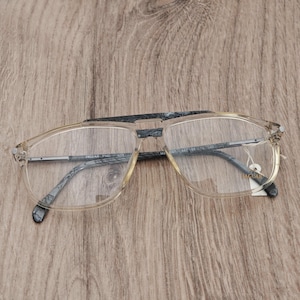May include: Jaguar brand spectacles featuring a clear, transparent frame with a distinctive grey and black marbled pattern on the top brow bar and temples. The lenses are clear, and the word Jaguar is printed in gold on the right lens.