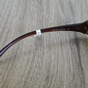 May include: Close-up of a pair of brown sunglasses with a curved arm. The text "MOD. 8009 094/S81 120" is printed on the arm. The sunglasses are on a wooden surface.