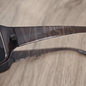 Puede incluir: Gafas de sol de color gris oscuro transl&uacute;cido con la palabra VERSUS grabada en letras may&uacute;sculas grandes a lo largo del lado de la patilla. La montura est&aacute; fabricada con un material liso y semitransparente que deja pasar la luz.