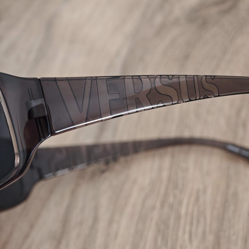 Puede incluir: Gafas de sol de color gris oscuro transl&uacute;cido con la palabra VERSUS grabada en letras may&uacute;sculas grandes a lo largo del lado de la patilla. La montura est&aacute; fabricada con un material liso y semitransparente que deja pasar la luz.