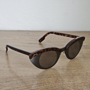 Puede incluir: Gafas de sol de ojo de gato con montura y patillas con estampado de carey. Las lentes son de color gris oscuro. Las gafas de sol están sobre una superficie de madera marrón claro.