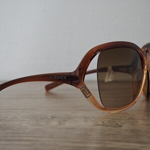 Puede incluir: Gafas de sol Versace marrones con tinte degradado. Las gafas tienen una montura gruesa con el logo de Versace en la patilla. Las lentes son de color marr&oacute;n oscuro y la montura es de color marr&oacute;n claro.