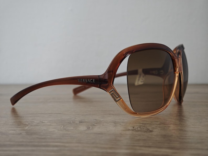 Puede incluir: Gafas de sol Versace marrones con tinte degradado. Las gafas tienen una montura gruesa con el logo de Versace en la patilla. Las lentes son de color marr&oacute;n oscuro y la montura es de color marr&oacute;n claro.