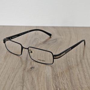 Peut inclure: Lunettes Prada rectangulaires noires avec verres transparents. Les montures sont noires avec des accents argentés sur les branches. Le logo Prada est visible sur la lentille droite. Les lunettes sont sur une surface en bois.