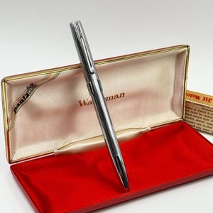 K&ouml;nnte beinhalten: Ein silberner Waterman-Stift in einer offenen, rot samtgef&uuml;tterten Schachtel. Der Stift ist abgewinkelt, um sein schlankes Design hervorzuheben. Die Schachtel hat einen goldenen Rand und das Wort "Waterman" ist sichtbar.