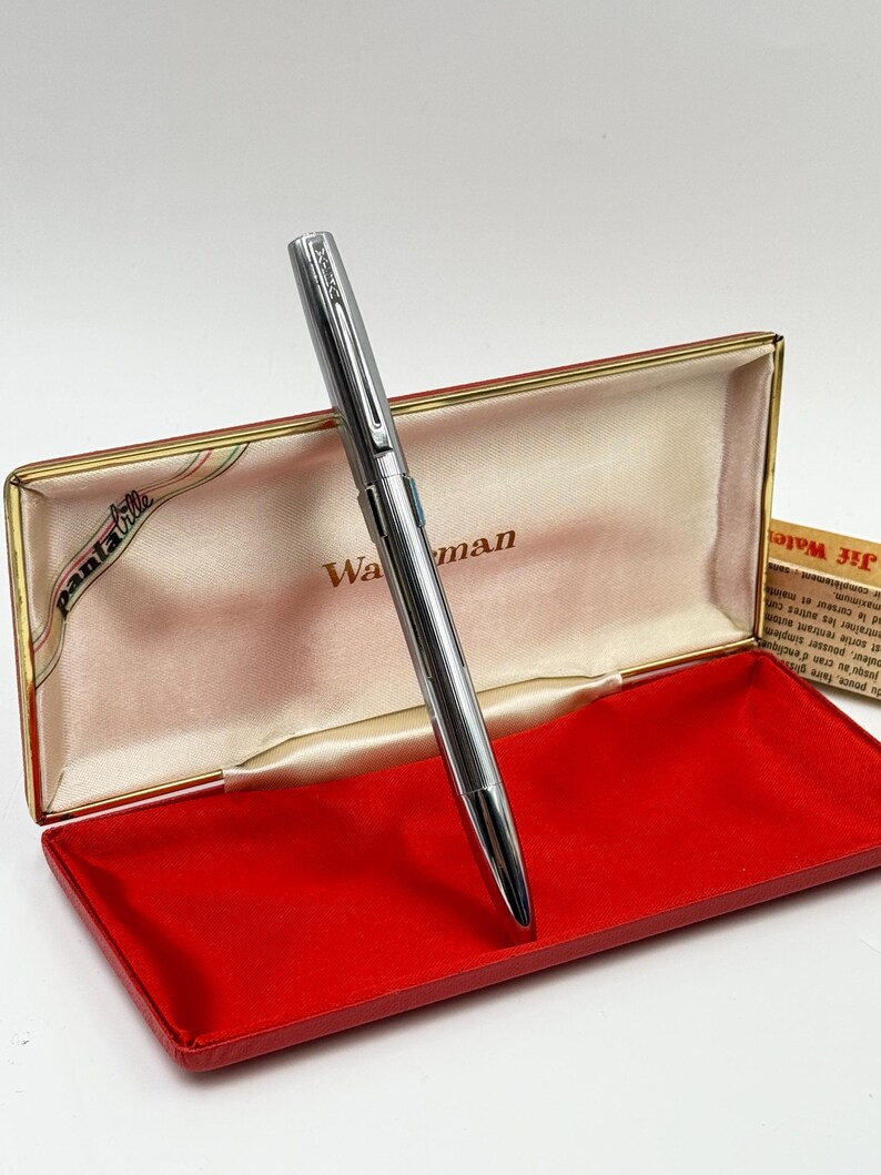 K&ouml;nnte beinhalten: Ein silberner Waterman-Stift in einer offenen, rot samtgef&uuml;tterten Schachtel. Der Stift ist abgewinkelt, um sein schlankes Design hervorzuheben. Die Schachtel hat einen goldenen Rand und das Wort "Waterman" ist sichtbar.