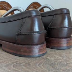 May include: Close-up of a pair of brown leather loafers. The shoes have a classic design with a polished finish. The soles are a lighter brown, and the heels show wear. Shoe trees are inserted in the shoes.