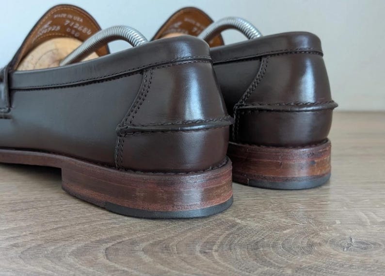 May include: Close-up of a pair of brown leather loafers. The shoes have a classic design with a polished finish. The soles are a lighter brown, and the heels show wear. Shoe trees are inserted in the shoes.