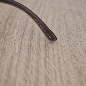 Puede incluir: Una patilla de gafas de color marr&oacute;n oscuro y transl&uacute;cido. En la parte interior de la patilla hay un texto impreso en blanco que dice VERSUS, MADE IN ITALY y el marcado CE, indicando el origen y la conformidad de la montura de las gafas.