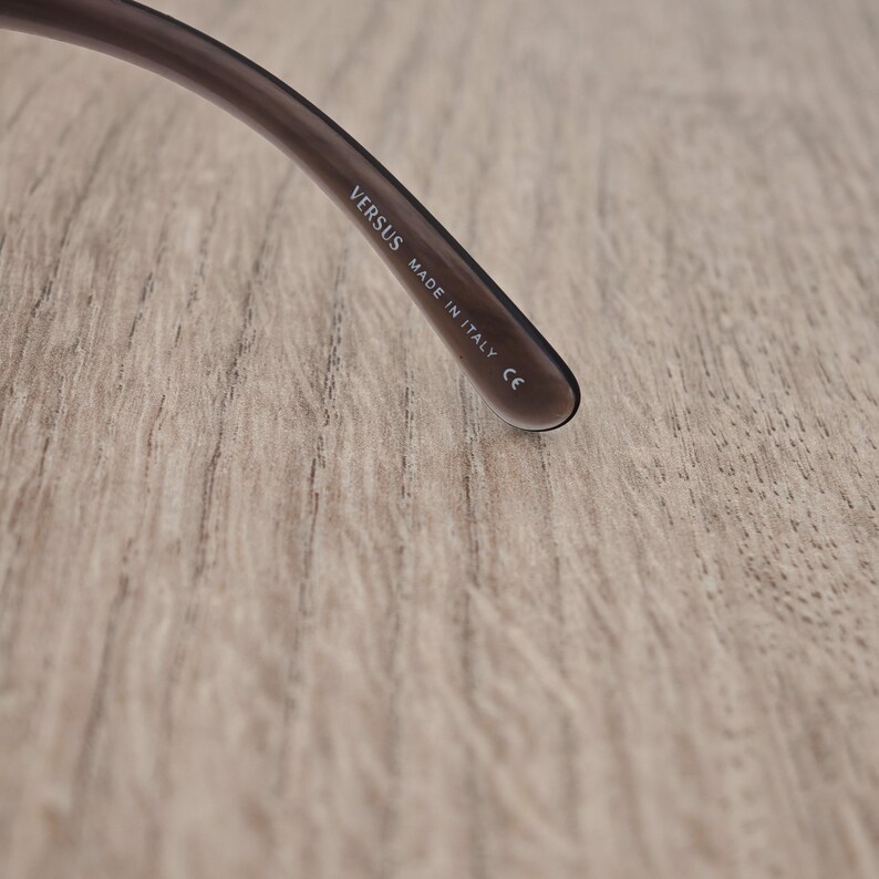 Puede incluir: Una patilla de gafas de color marr&oacute;n oscuro y transl&uacute;cido. En la parte interior de la patilla hay un texto impreso en blanco que dice VERSUS, MADE IN ITALY y el marcado CE, indicando el origen y la conformidad de la montura de las gafas.