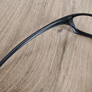 May include: Black eyeglasses with a sleek, modern design. The frames are a solid black color, with a curved temple arm extending from the lens frame. The glasses are set against a light brown wooden surface.