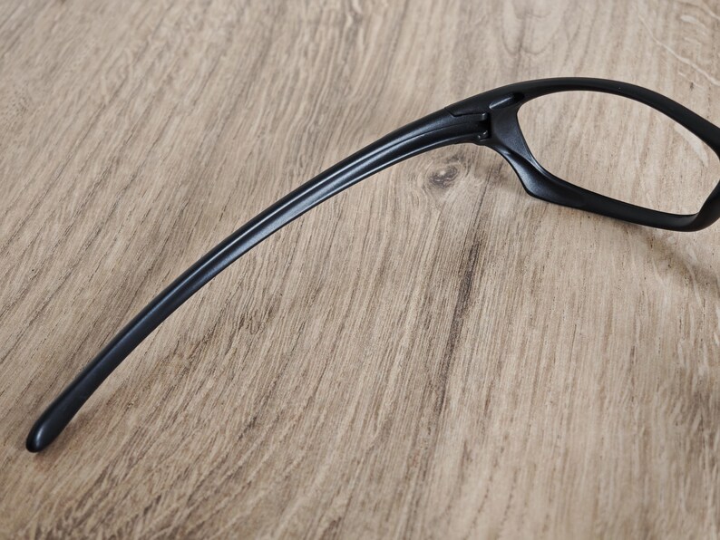 May include: Black eyeglasses with a sleek, modern design. The frames are a solid black color, with a curved temple arm extending from the lens frame. The glasses are set against a light brown wooden surface.
