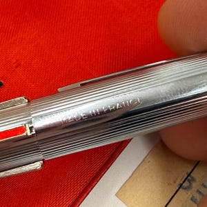 K&ouml;nnte beinhalten: Nahaufnahme eines silbernen Stifts mit geripptem Design. Der Stift hat einen roten Akzent und die Worte "MADE IN FRANCE" sind auf der Seite eingraviert. Der Stift liegt auf einer roten Oberfl&auml;che.