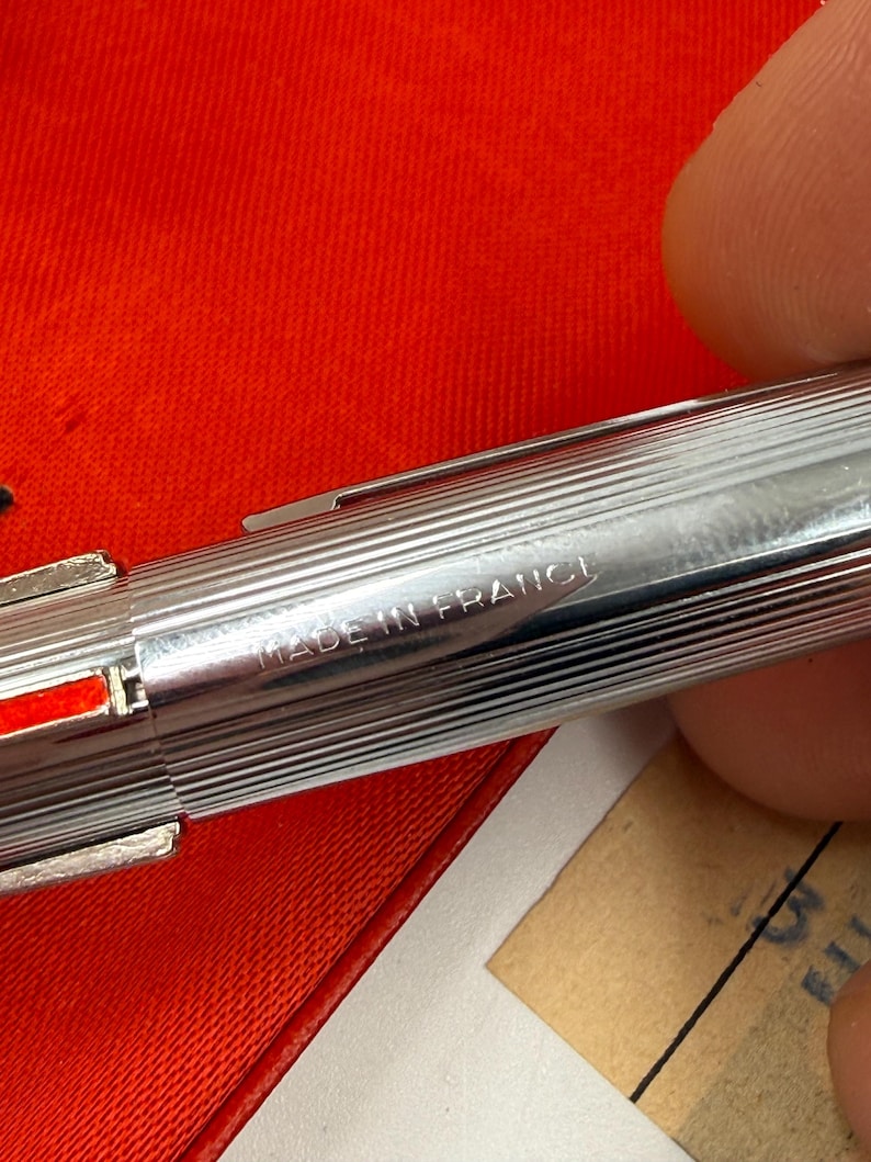 K&ouml;nnte beinhalten: Nahaufnahme eines silbernen Stifts mit geripptem Design. Der Stift hat einen roten Akzent und die Worte "MADE IN FRANCE" sind auf der Seite eingraviert. Der Stift liegt auf einer roten Oberfl&auml;che.