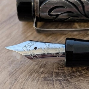 以下が含まれることがあります： 銀色のペン先と黒い胴体の万年筆のクローズアップ。ペン先には複雑な彫刻模様と「Lapis Iridium」の文字が刻まれています。ペンの胴体には装飾的な花の模様があります。