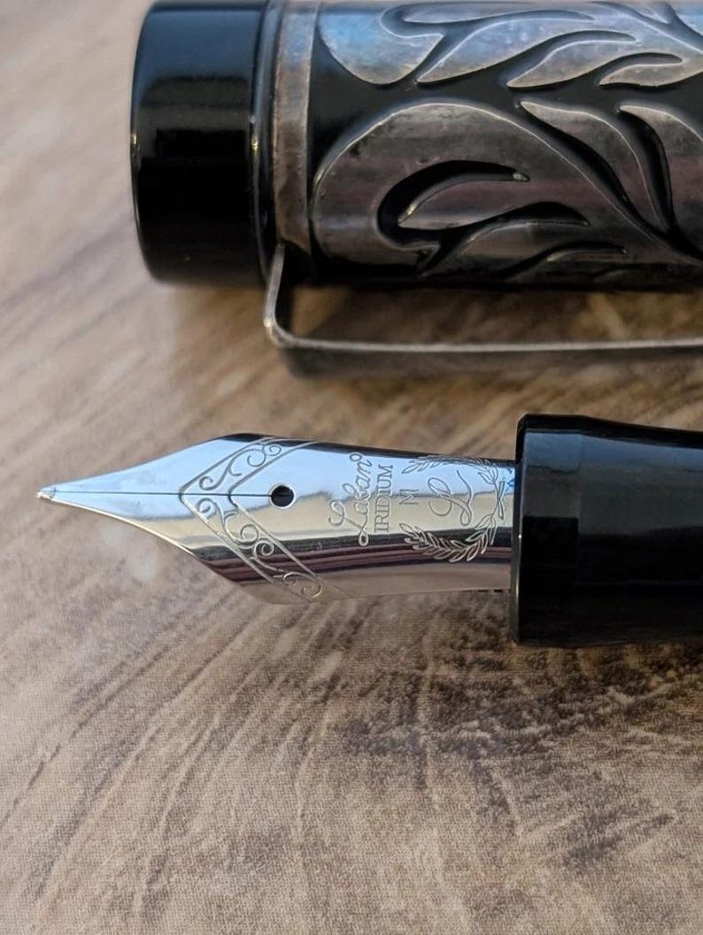 以下が含まれることがあります： 銀色のペン先と黒い胴体の万年筆のクローズアップ。ペン先には複雑な彫刻模様と「Lapis Iridium」の文字が刻まれています。ペンの胴体には装飾的な花の模様があります。