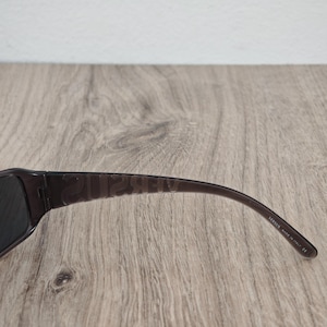 Puede incluir: Gafas de sol de color marr&oacute;n oscuro con montura transl&uacute;cida y la palabra VERSUS grabada en letras may&uacute;sculas grandes a lo largo de la patilla. El interior de la patilla muestra el texto blanco VERSUS MADE IN ITALY CE cerca de la curva.