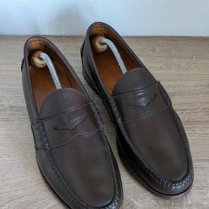 May include: A pair of dark brown leather penny loafers with a classic design. The shoes have a rounded toe, a penny strap across the vamp, and a stitched sole. Shoe trees are inserted inside the shoes. The loafers are displayed on a light brown wooden surface.