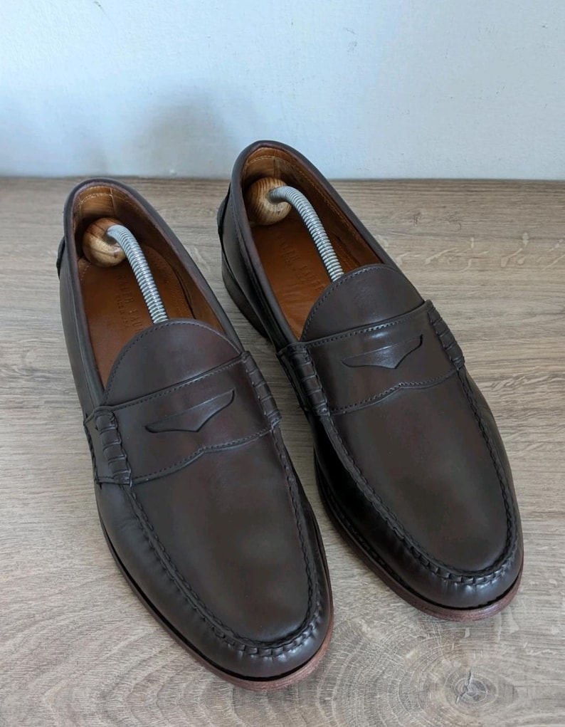 May include: A pair of dark brown leather penny loafers with a classic design. The shoes have a rounded toe, a penny strap across the vamp, and a stitched sole. Shoe trees are inserted inside the shoes. The loafers are displayed on a light brown wooden surface.