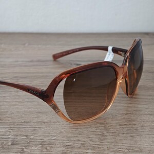 Puede incluir: Gafas de sol Versace marrones con lentes degradadas. Las monturas son una mezcla de marr&oacute;n claro y oscuro, con el nombre de la marca visible en la patilla. Las gafas de sol est&aacute;n sobre una superficie de madera.