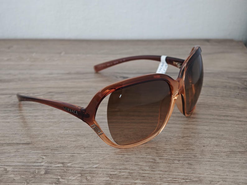 Puede incluir: Gafas de sol Versace marrones con lentes degradadas. Las monturas son una mezcla de marr&oacute;n claro y oscuro, con el nombre de la marca visible en la patilla. Las gafas de sol est&aacute;n sobre una superficie de madera.