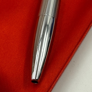 K&ouml;nnte beinhalten: Nahaufnahme eines silbernen Stifts mit geripptem Design auf der oberen H&auml;lfte. Der Stift liegt auf einer leuchtend roten Oberfl&auml;che, was einen auff&auml;lligen Kontrast erzeugt. Die Spitze des Stifts ist sichtbar, und die Gesamt&auml;sthetik ist elegant und modern.
