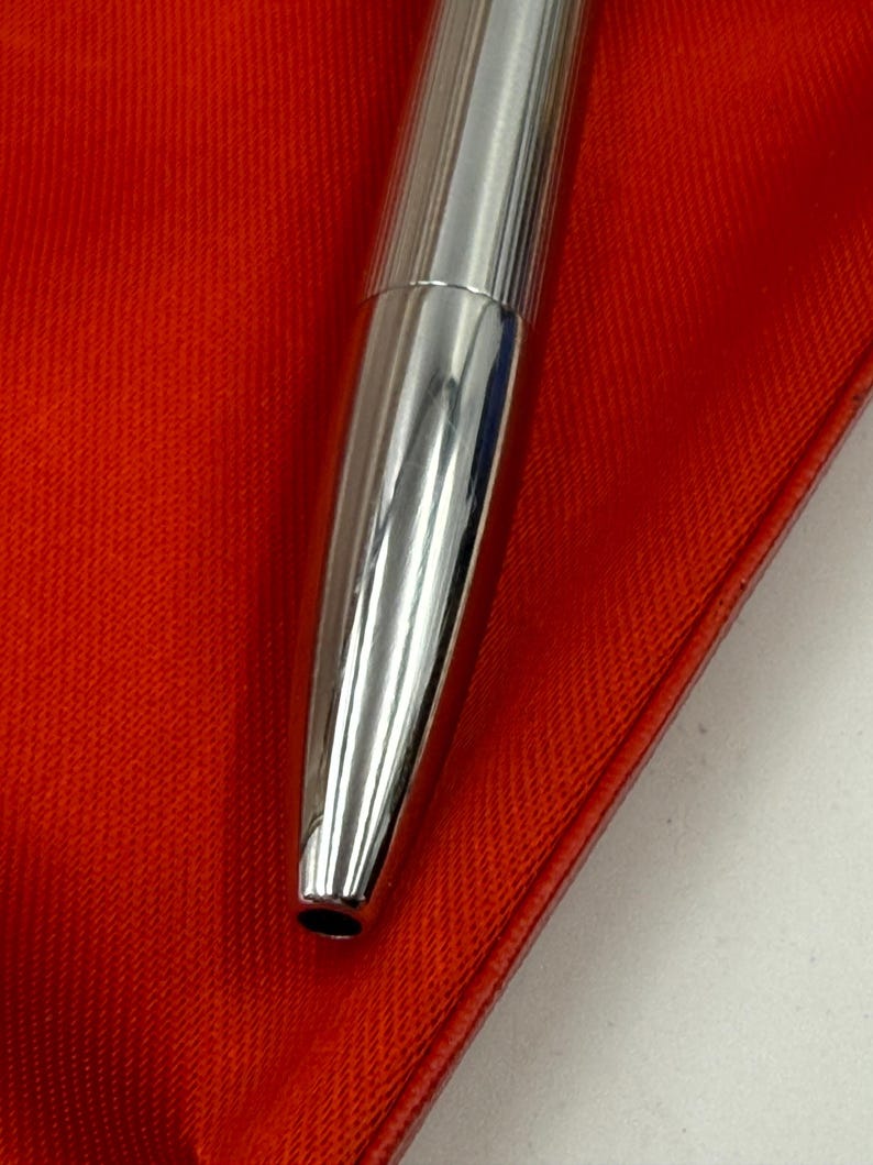 K&ouml;nnte beinhalten: Nahaufnahme eines silbernen Stifts mit geripptem Design auf der oberen H&auml;lfte. Der Stift liegt auf einer leuchtend roten Oberfl&auml;che, was einen auff&auml;lligen Kontrast erzeugt. Die Spitze des Stifts ist sichtbar, und die Gesamt&auml;sthetik ist elegant und modern.