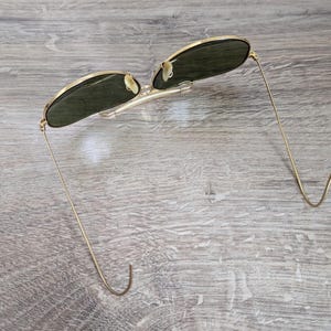 K&ouml;nnte beinhalten: Sonnenbrille mit goldfarbenem Rahmen und dunkelgr&uuml;nen Gl&auml;sern. Die d&uuml;nnen, goldfarbenen B&uuml;gel sind an den Enden gebogen. Die Sonnenbrille liegt auf einer hellgrauen Holzoberfl&auml;che.