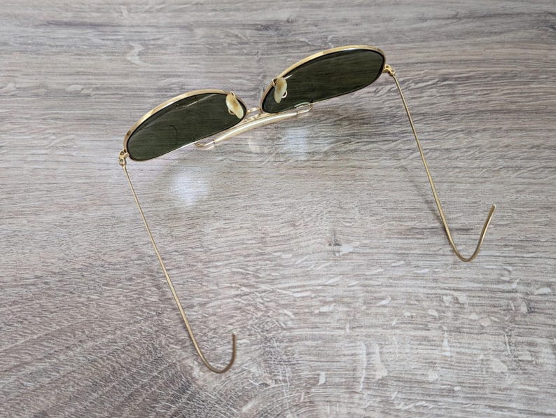 K&ouml;nnte beinhalten: Sonnenbrille mit goldfarbenem Rahmen und dunkelgr&uuml;nen Gl&auml;sern. Die d&uuml;nnen, goldfarbenen B&uuml;gel sind an den Enden gebogen. Die Sonnenbrille liegt auf einer hellgrauen Holzoberfl&auml;che.