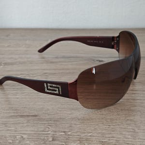 May include: Brown sunglasses with a gradient lens. The frames are a dark burgundy color with a decorative silver emblem on the temples. The sunglasses are resting on a wooden surface.