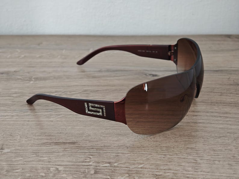 May include: Brown sunglasses with a gradient lens. The frames are a dark burgundy color with a decorative silver emblem on the temples. The sunglasses are resting on a wooden surface.