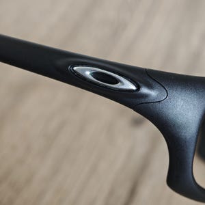 May include: Close-up of a black eyeglass frame with a silver oval logo on the temple. The frame has a sleek, modern design. The background is a light brown, out-of-focus surface.