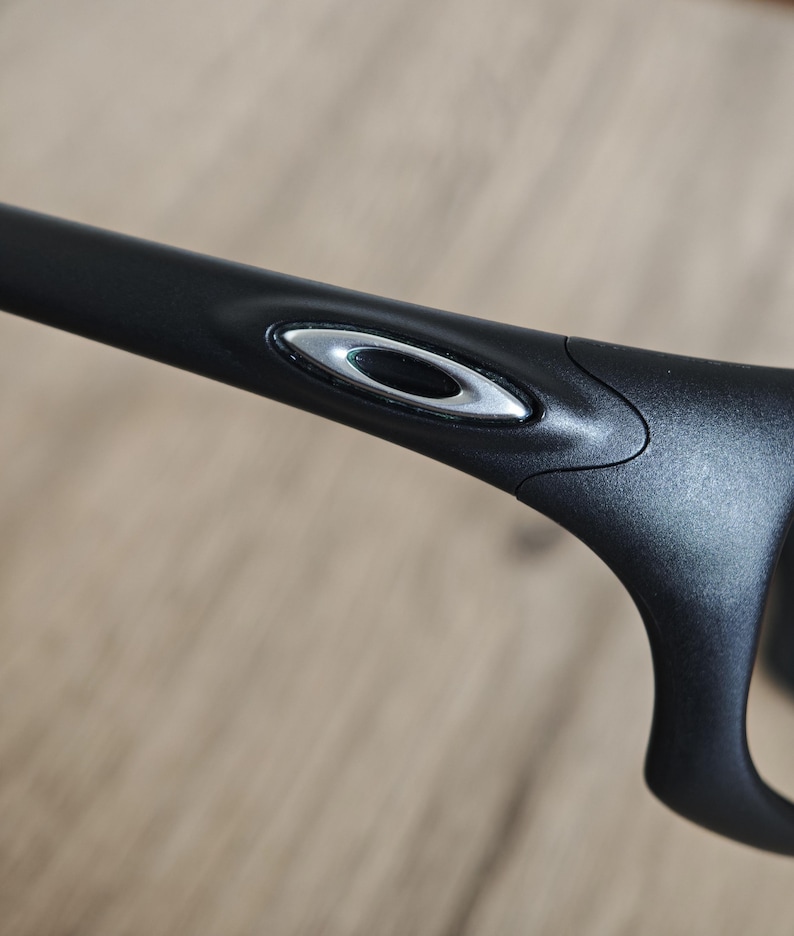 May include: Close-up of a black eyeglass frame with a silver oval logo on the temple. The frame has a sleek, modern design. The background is a light brown, out-of-focus surface.