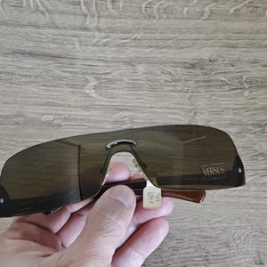 May include: Brown Versus sunglasses with a dark tint. The sunglasses have a curved, shield-like design with a silver bridge and brown arms. The Versus logo is visible on the right lens.
