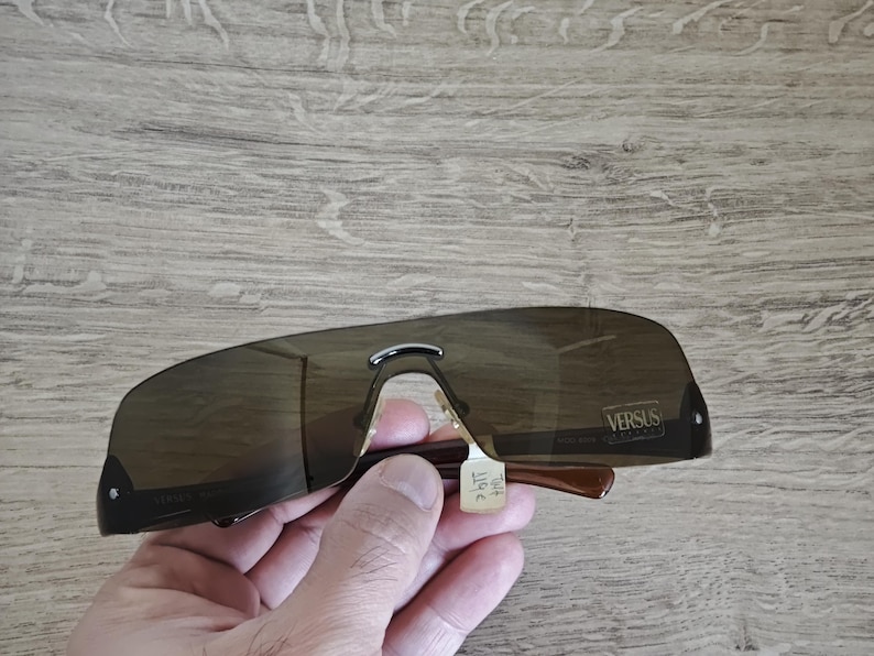May include: Brown Versus sunglasses with a dark tint. The sunglasses have a curved, shield-like design with a silver bridge and brown arms. The Versus logo is visible on the right lens.