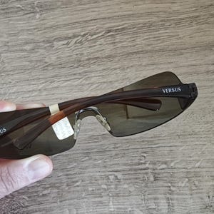 May include: Brown rimless sunglasses with dark lenses. The arms are brown with the word "VERSUS" printed in white on the temples. The sunglasses are held up, showing the back of the frame.