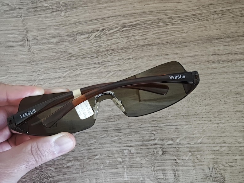 May include: Brown rimless sunglasses with dark lenses. The arms are brown with the word "VERSUS" printed in white on the temples. The sunglasses are held up, showing the back of the frame.