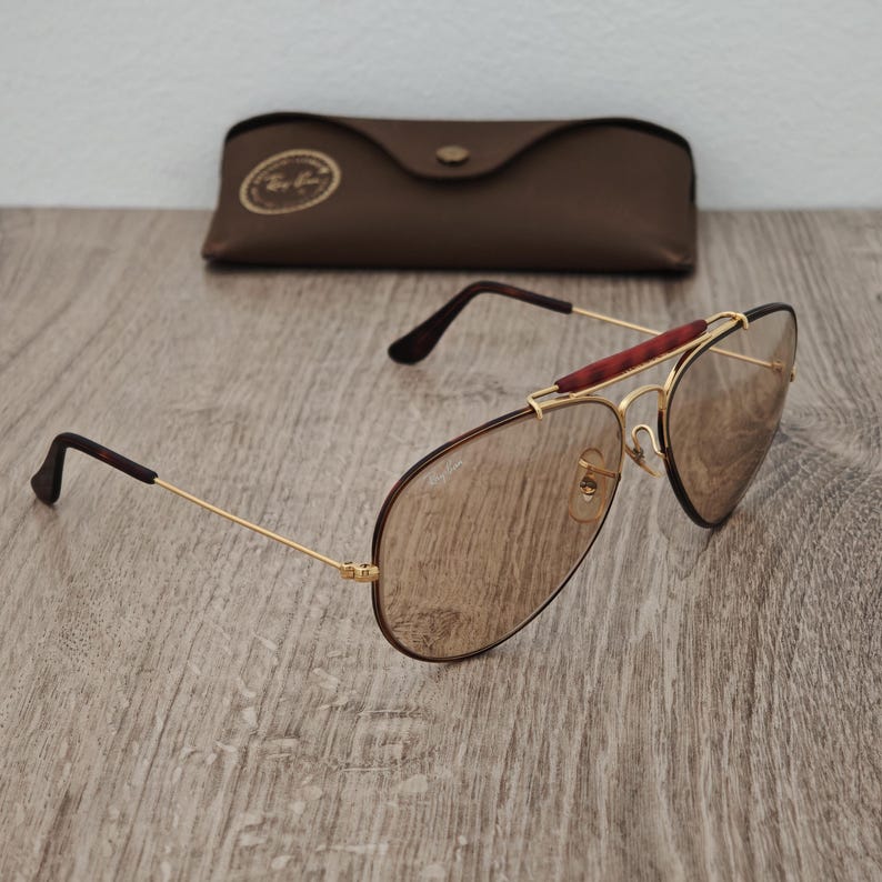 K&ouml;nnte beinhalten: Braune Aviator-Sonnenbrille mit goldfarbenem Rahmen und braunem Lederetui. Die Sonnenbrille hat einen Doppelsteg und einen braunen Akzent auf dem oberen B&uuml;gel. Die Gl&auml;ser sind hellbraun.