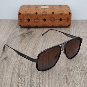 May include: Black aviator sunglasses with brown gradient lenses and a matching brown case. The sunglasses have a metal frame with a mesh detail on the bridge and the MCM logo on the temples. The case is brown with the MCM logo pattern.